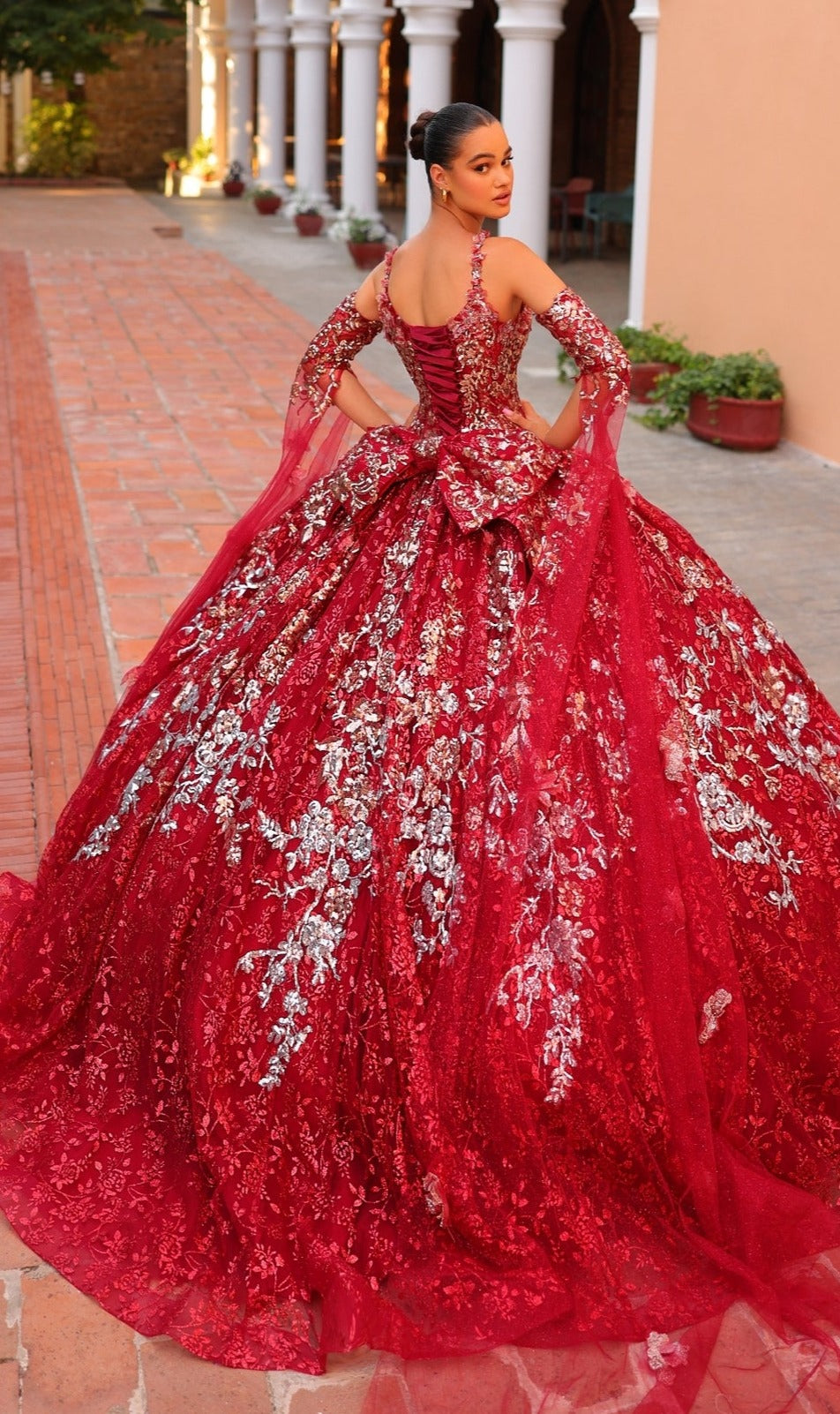 A woman wears the Quinceanera Dress 54322 by Amarra?an ornate, bright red gown with silver floral details?and stands on a tiled walkway, looking over her shoulder at the camera.